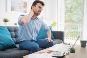 Man rubs his sore neck while sitting at makeshift home workstation, home office ergonomics